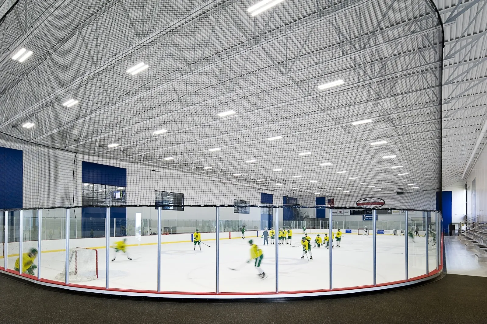 Jugadores repartidos por la pista vistos desde las gradas, con un largo techo blanco acanalado arriba y vallas y vidrio azules alrededor del hielo. — Prairie Lakes Ice Arena