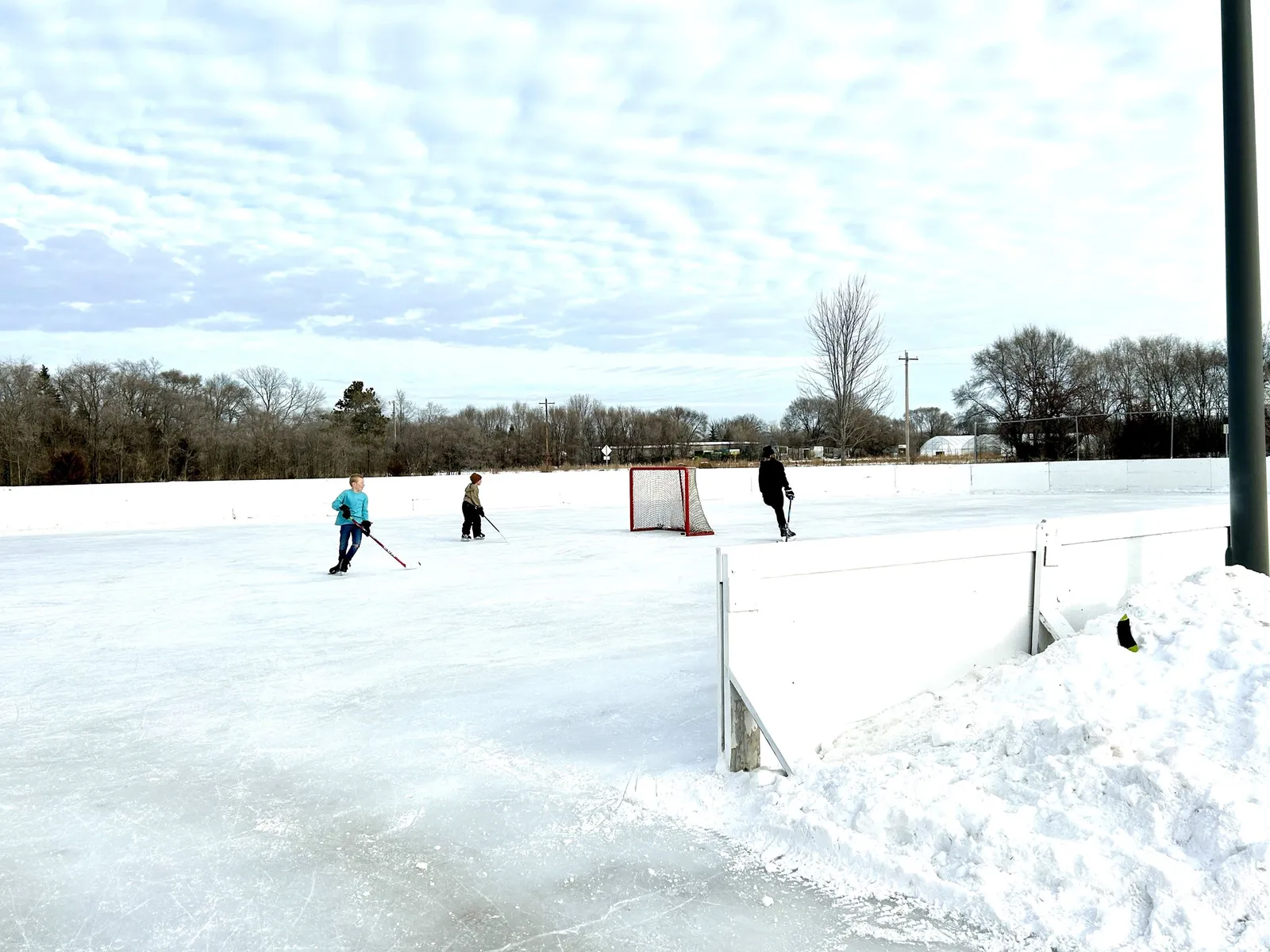 The outdoor hockey rink at Prairie Park in Otsego in winter, with a net and a few players at a distance on the snow-banked ice. — Prairie Park Rinks