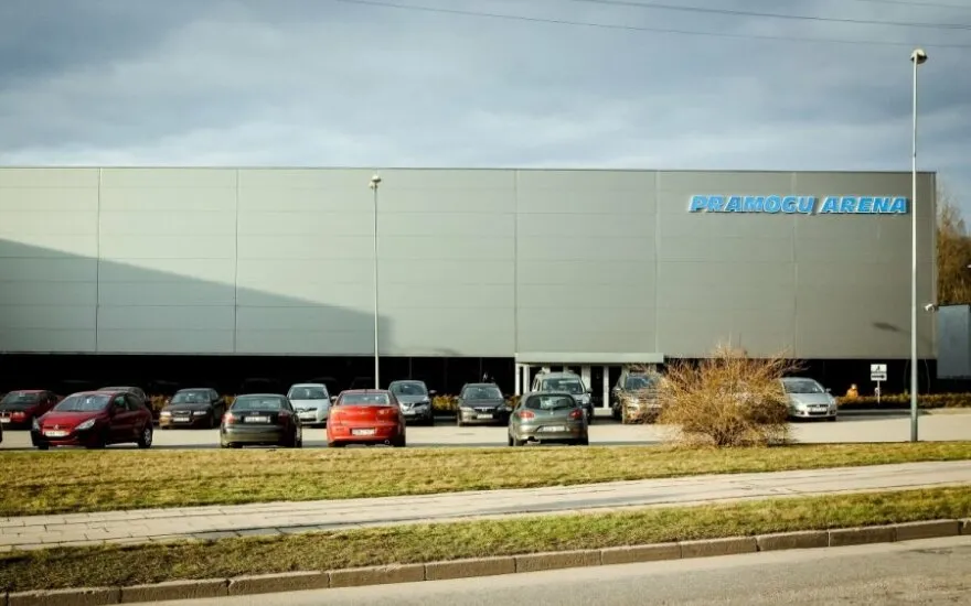 Ilga pilka Pramogų arenos šoninė siena su mėlynais užrašais, matoma per Ąžuolyno gatvės automobilių stovėjimo aikštelę. – Pramogų Arena