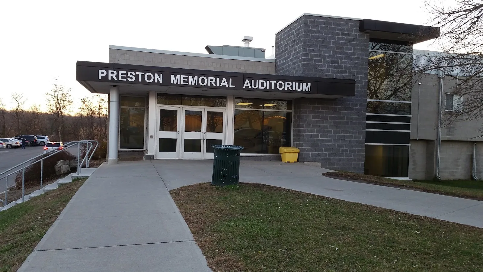 La entrada del Preston Memorial Auditorium, con su nombre sobre una marquesina oscura encima de puertas de vidrio, junto a una torre de bloques gris y con un andador al frente. — Preston Memorial Auditorium