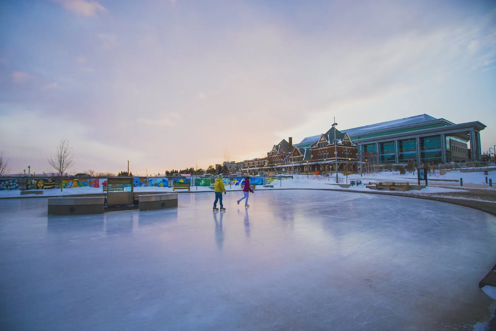 La pista refrigerada de Prince Arthur's Landing al atardecer, con dos patinadores en el hielo y el pabellón junto al agua más allá — Prince Arthur's Landing Ice Rink