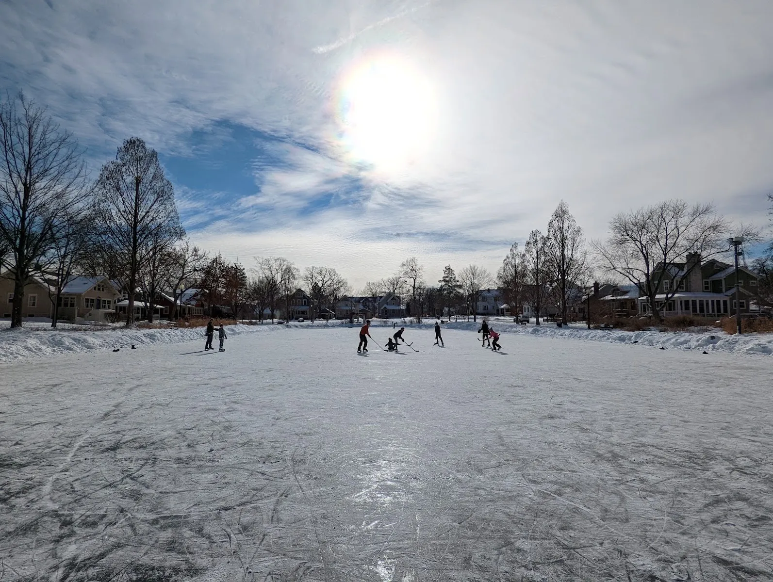 Skaters playing hockey on the frozen pond in low winter sun, houses beyond — Prince Pond Ice Rink