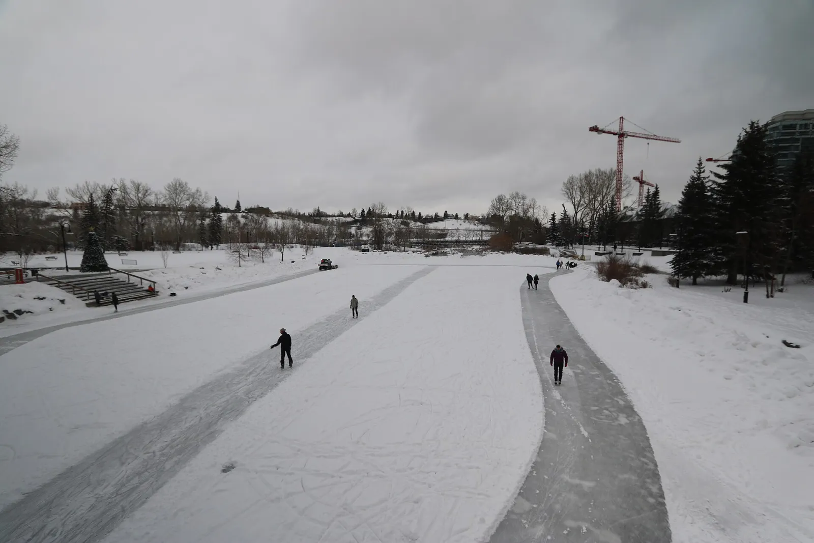 Dos patinadores en el camino de hielo despejado de la laguna de Prince's Island en invierno, con una orilla nevada y una grúa de construcción más allá — Prince's Island Park Outdoor Skating Rink