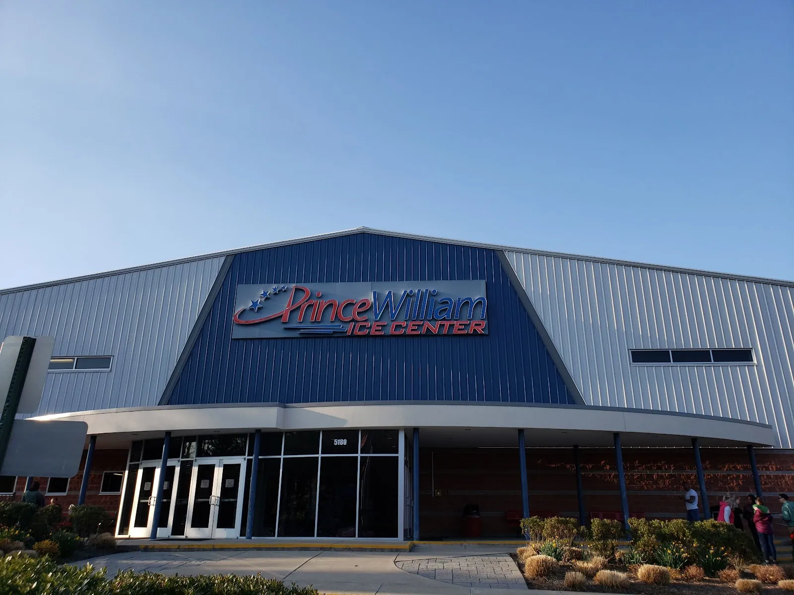 La fachada de Prince William Ice Center, con su nombre en un panel azul sobre la marquesina curva de vidrio de la entrada y una base de ladrillo. — Prince William Ice Center