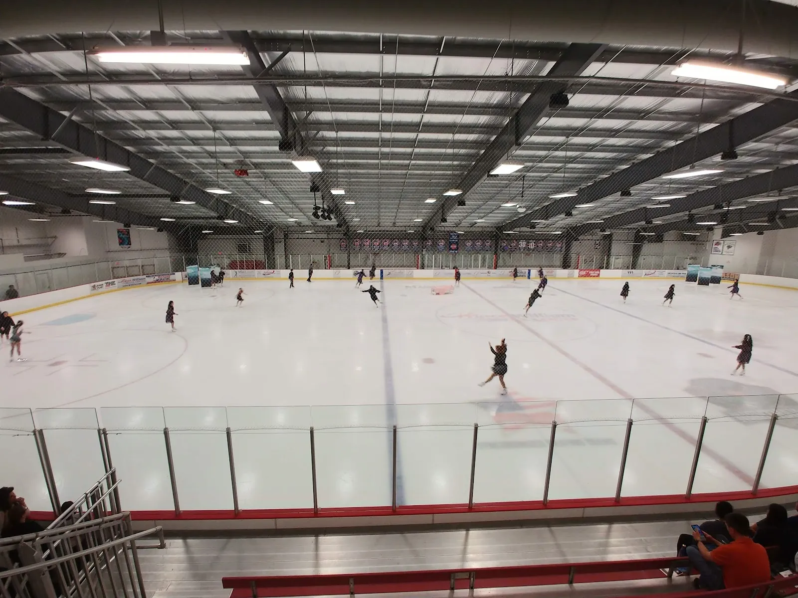 Una vista amplia de la pista desde las gradas, con jugadores repartidos por el hielo y bancas y vidrio a lo largo del lado cercano bajo un techo oscuro. — Prince William Ice Center