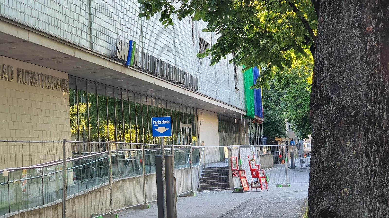 Die geflieste Front des Stadions mit dem verglasten Foyer und dem blauen Schriftzug neben einem Baumstamm, mit einer Rampe und Geländern, die zum Eingang führen. – Prinzregentenstadion Eislaufbahn