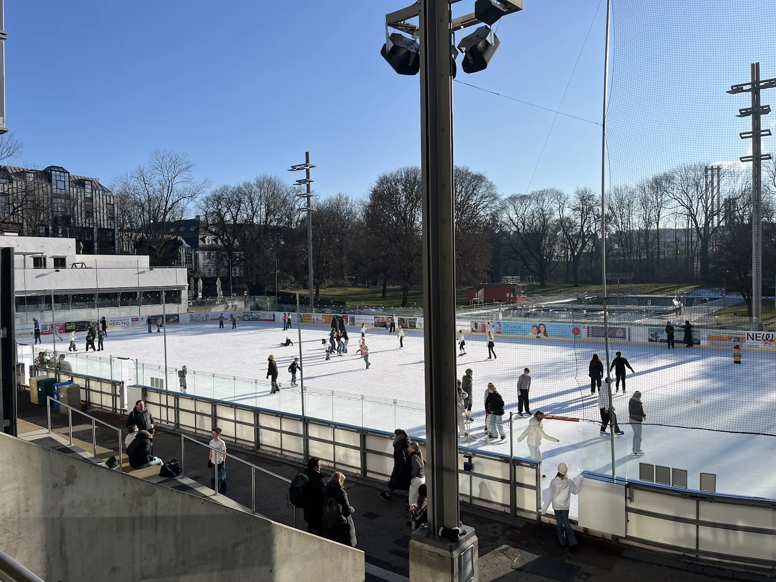 Die Freiluft-Eisbahn in der Wintersonne, eine weite weiße Eisfläche mit Läufern, Flutlichtmasten und den niedrigen Gebäuden des Stadions und kahlen Bäumen hinter der Bande. – Prinzregentenstadion Eislaufbahn