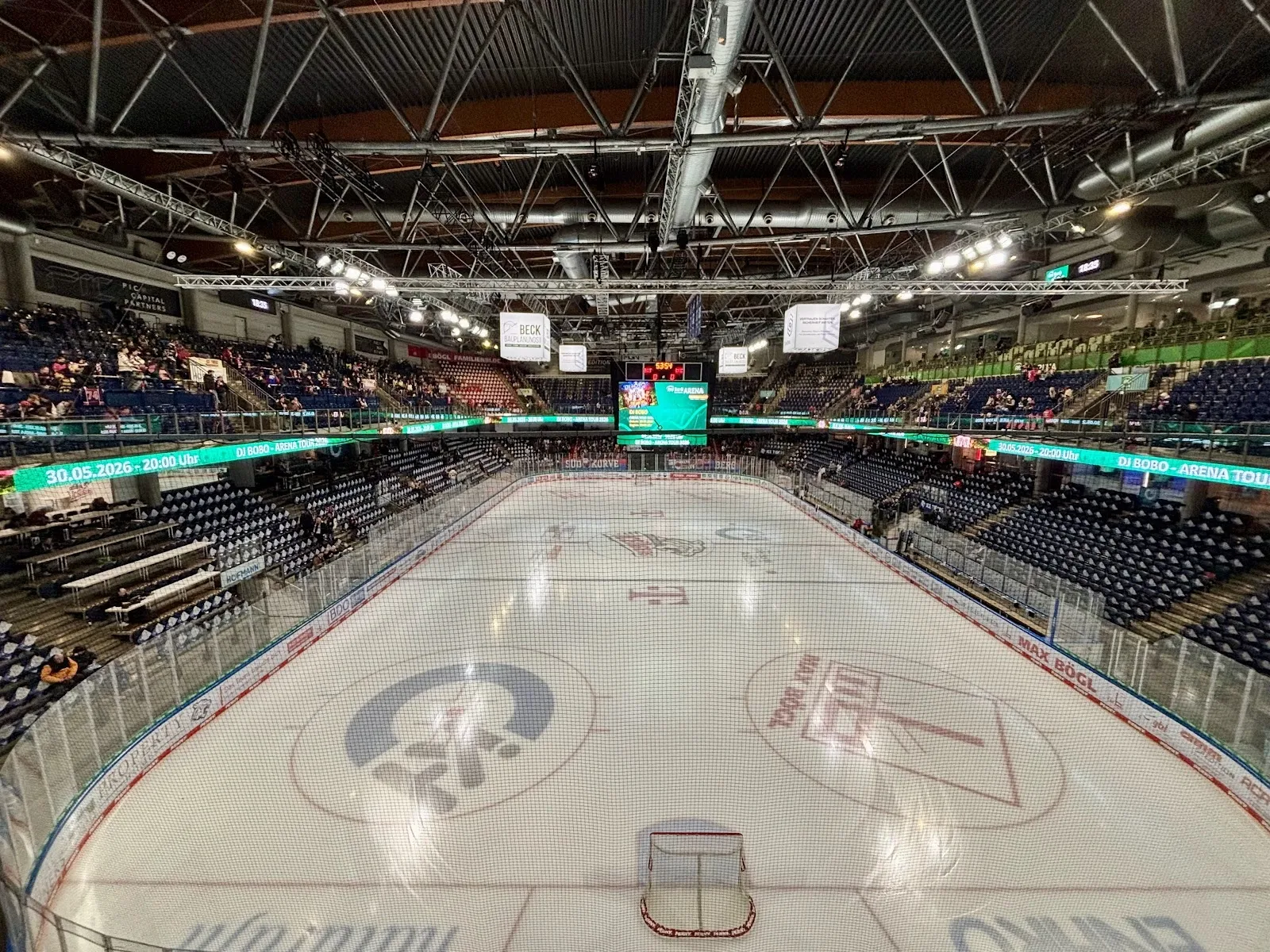 The empty ice sheet of the PSD Bank Nürnberg Arena seen from the stands, with the centre-ice and Ice Tigers markings, the scoreboard cube and the tiers of seating. — PSD Bank Nürnberg ARENA
