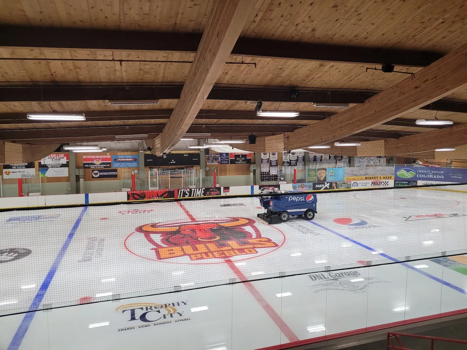 Toda la pista desde las gradas, con el logotipo rojo de los Pueblo Bulls en el centro de la pista, vallas cubiertas de anuncios de patrocinadores locales, techo de vigas de madera y una pulidora de hielo estacionada; no hay personas en el hielo. — Pueblo Ice Arena