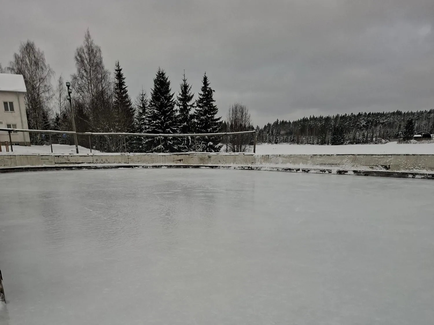 The glassy ice of the Pulkonkoski school rink inside its low concrete edge, with a white house and spruces behind under a grey sky. — Pulkonkosken koulun luistelukenttä