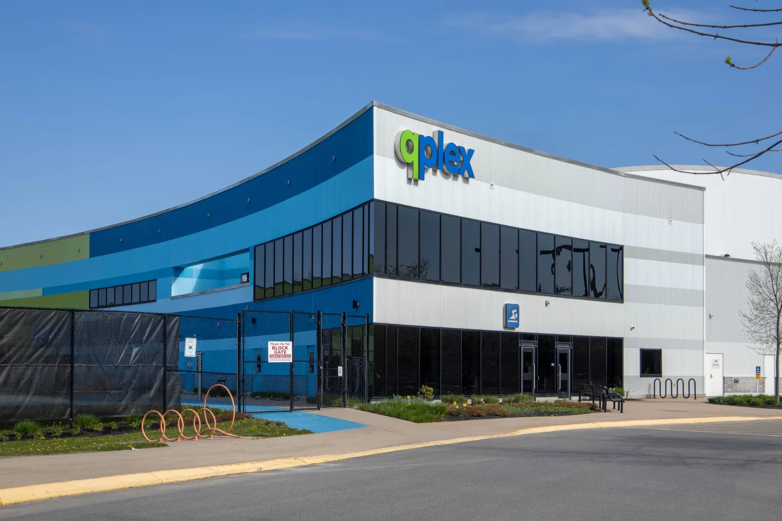 The qplex's curved, blue-and-white clad front with the green-and-blue qplex sign above dark glass entrance doors, on a sunny spring day. — Qplex