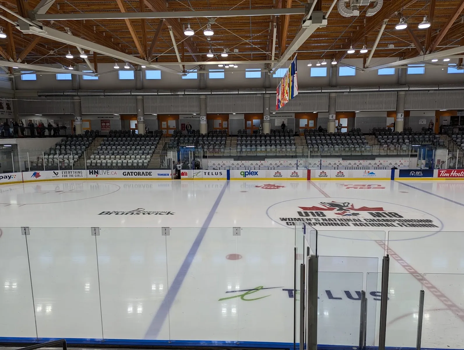 The NHL-size sheet from behind the glass, with the U18 Women's National Championship logo at centre ice, rows of grey seats and a timber roof. — Qplex