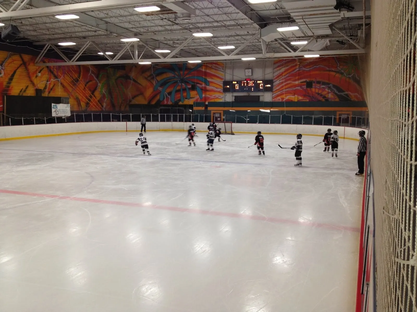 Un partido visto desde las gradas a través de una pista amplia, con jugadores repartidos por el hielo blanco debajo de un largo mural de hojas naranjas y rojas en la pared del fondo. — Queen City Sportsplex