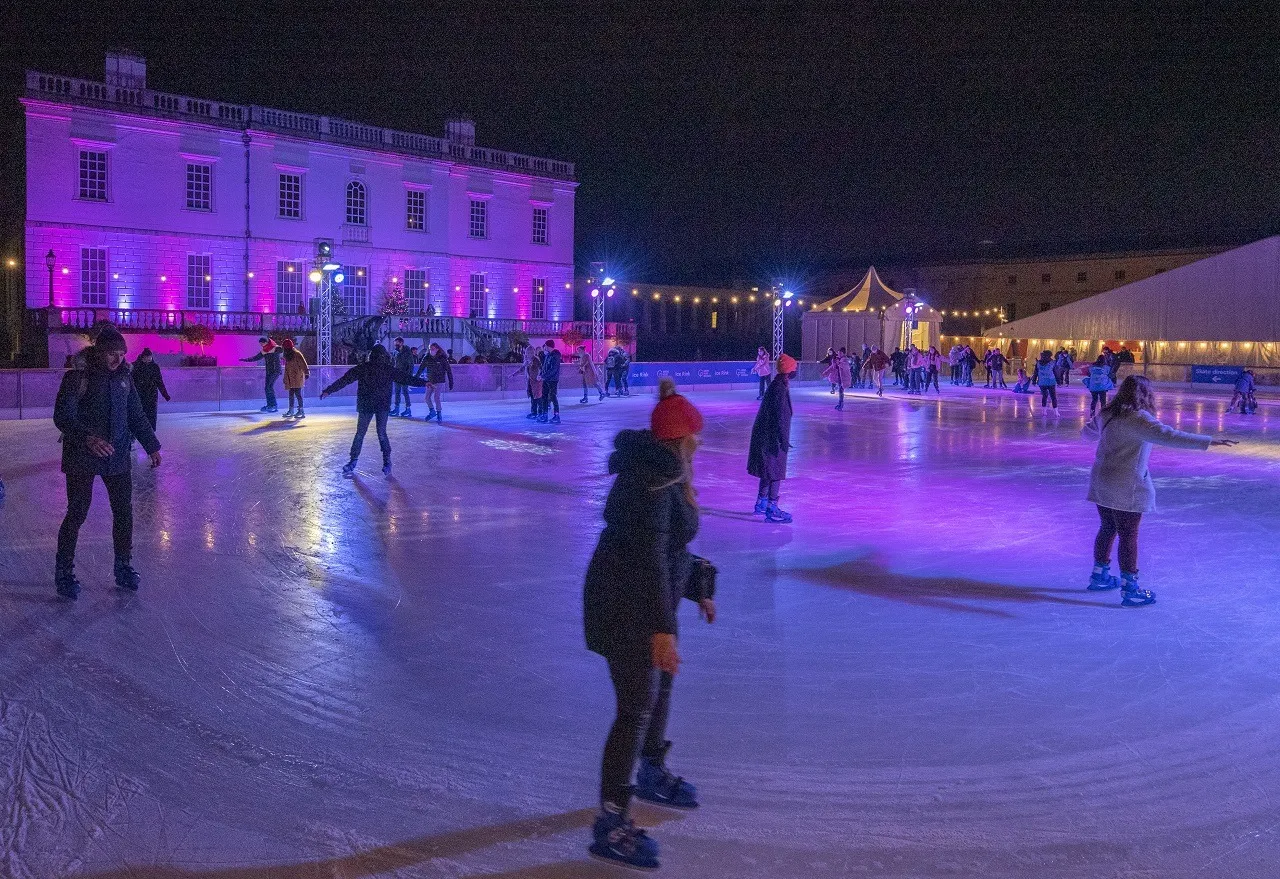 Skaters on the lit ice of the Queen's House rink in Greenwich at night, with the pink-lit white facade of the house behind. — Queen's House Ice Rink