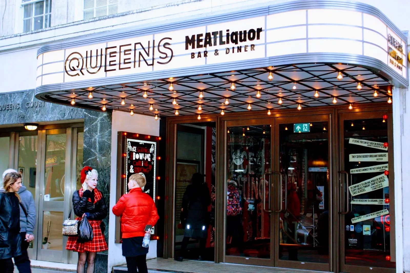 The street front of Queens on Queensway: a white canopy lined with bulbs reading QUEENS MEATLiquor Bar & Diner above the glass entrance doors, with passers-by at the door. — Queens Ice & Bowl