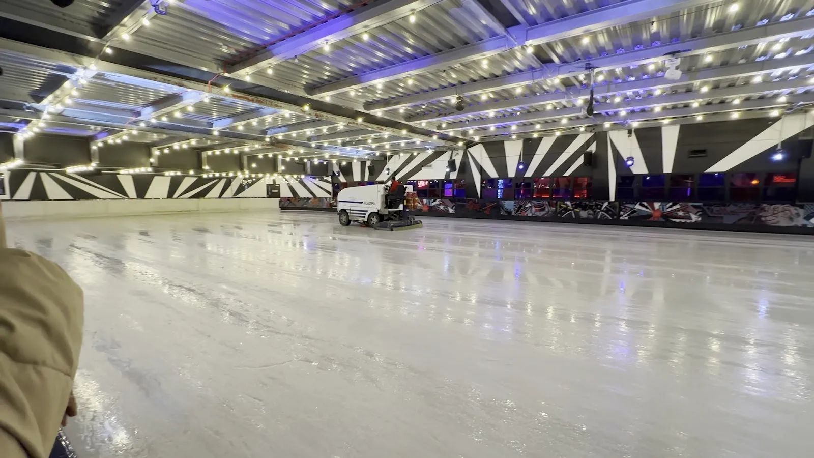 The empty ice sheet in the basement hall, resurfaced, with the ice-resurfacing machine at the far side and the striped black-and-white wall and string lights around the edge. — Queens Ice & Bowl