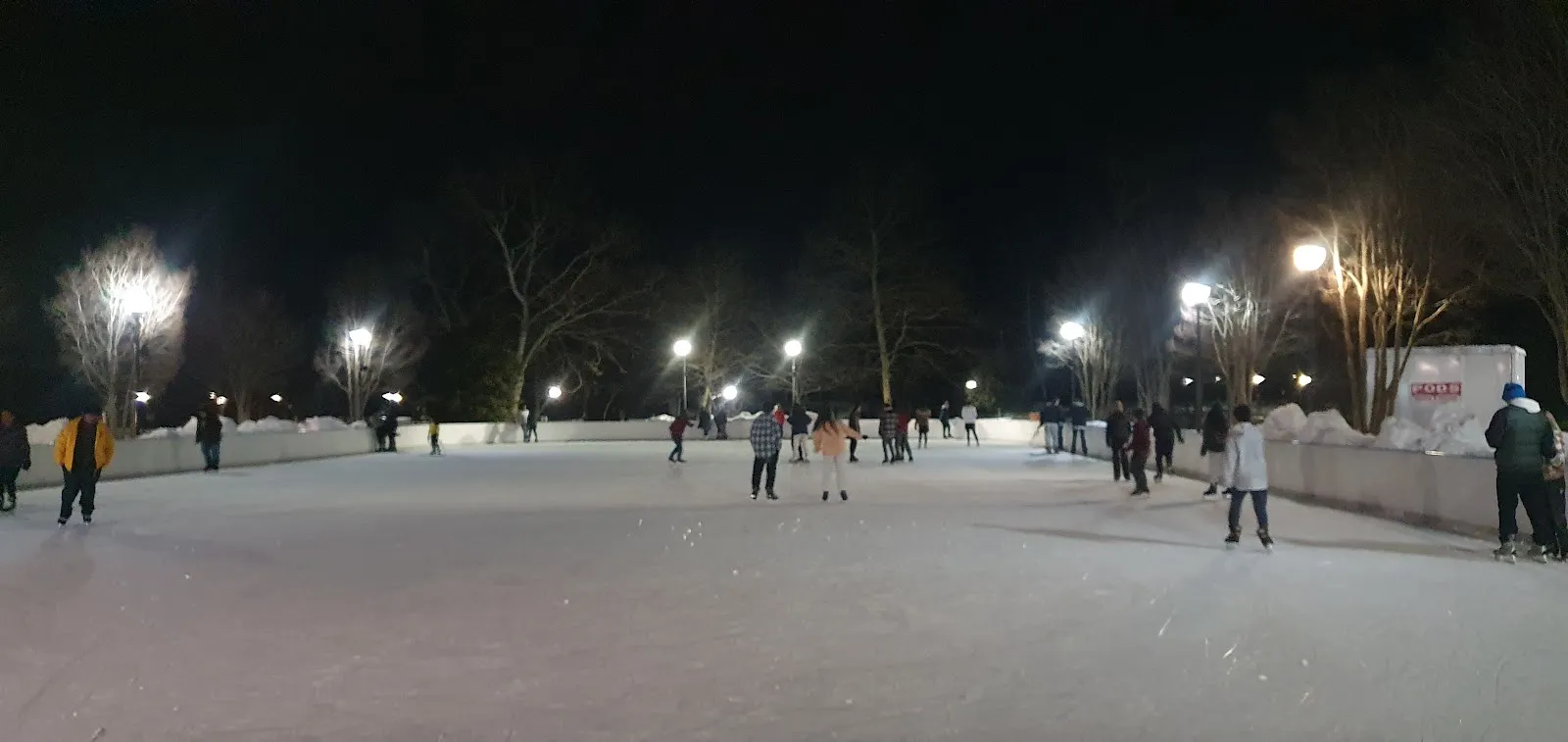 La pista exterior de noche desde el borde de la pista, con patinadores, un montón de nieve sobre las bandas y las luces del parque al fondo. — Quiet Waters Park Ice Rink