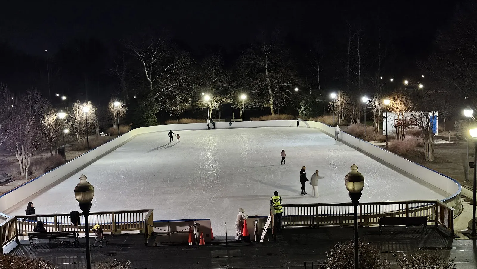 La pista exterior refrigerada de noche, vista desde la terraza de arriba, con algunos patinadores en el hielo y las luces del parque detrás. — Quiet Waters Park Ice Rink