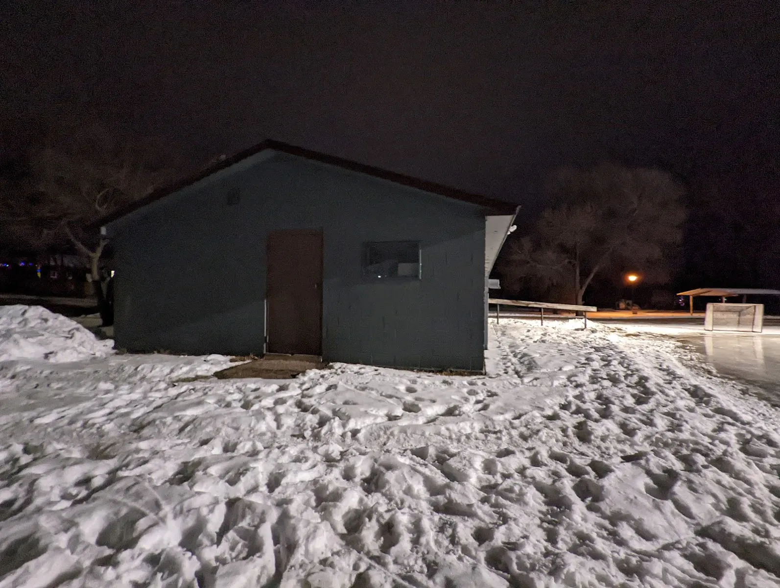 The park's blue warming house at night, with snow trampled in front of it, a lamp and the rink's boards and goal beyond. — Quincy Park Ice Rink