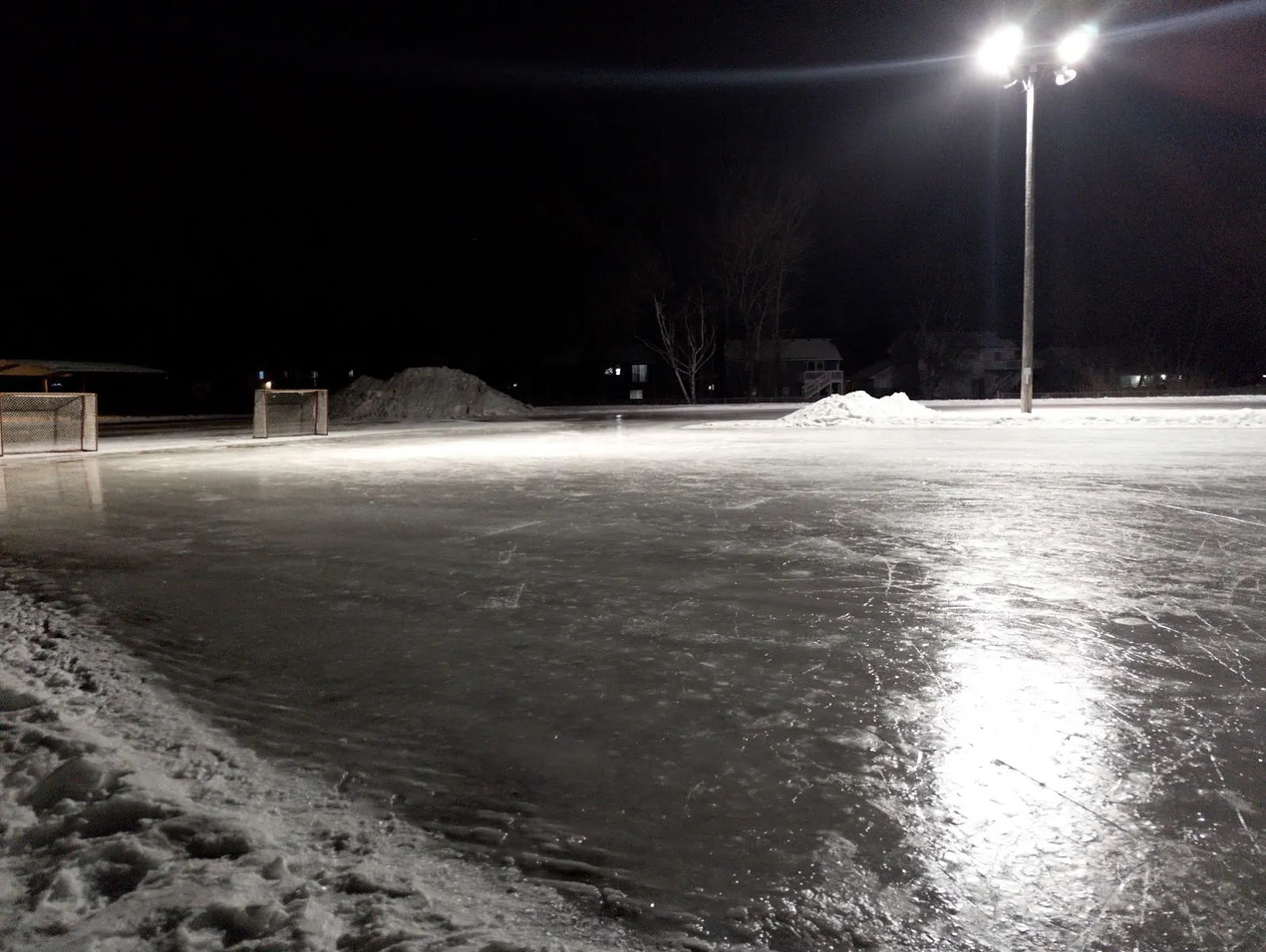 The outdoor rink at Quincy Park under floodlights at night, with glassy ice, snow banks along the edge and goals and shelters in the distance; no people. — Quincy Park Ice Rink