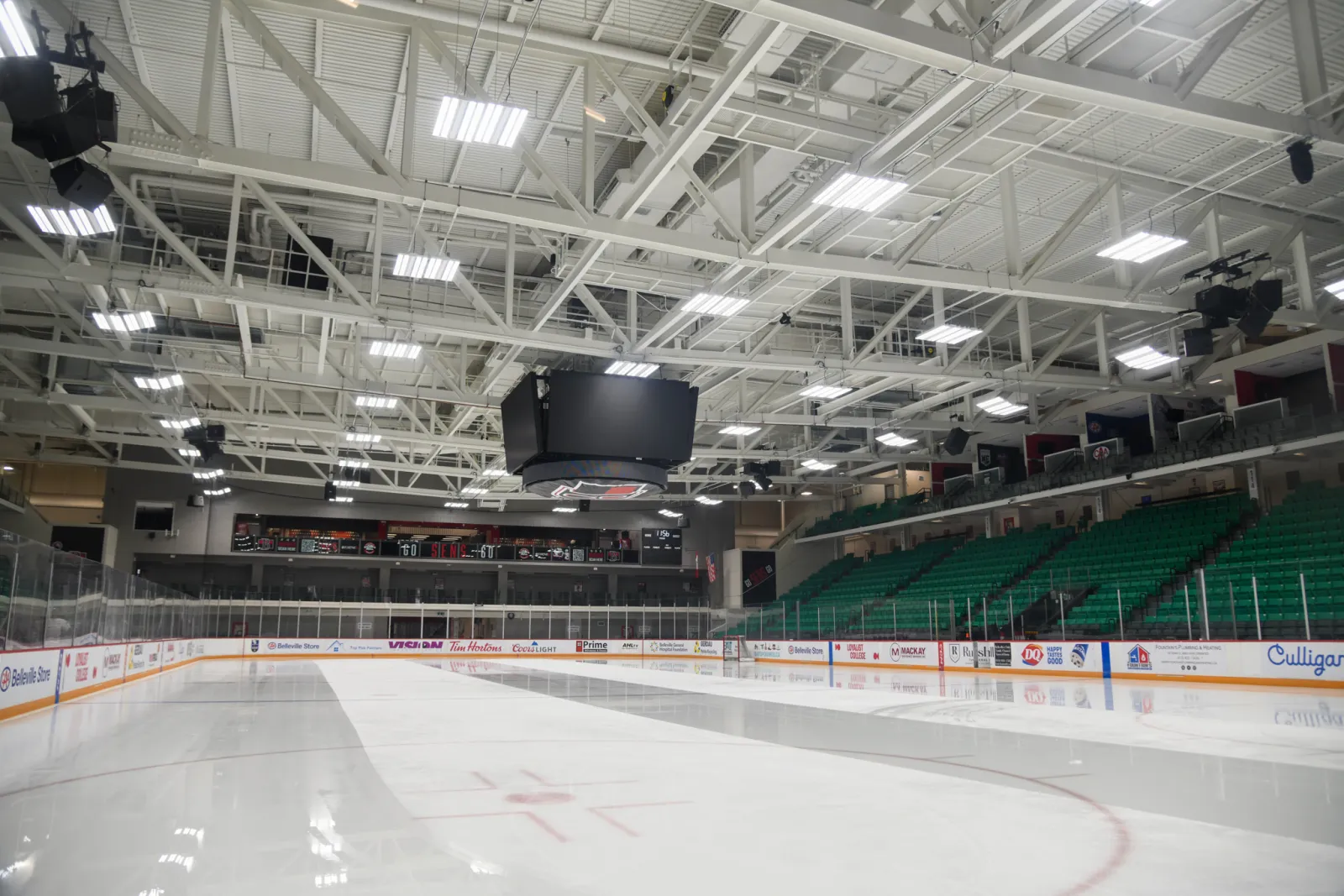The CAA Arena ice pad at the Quinte Sports & Wellness Centre in Belleville, with green seating along one side and a centre-hung scoreboard — Quinte Sports & Wellness Centre