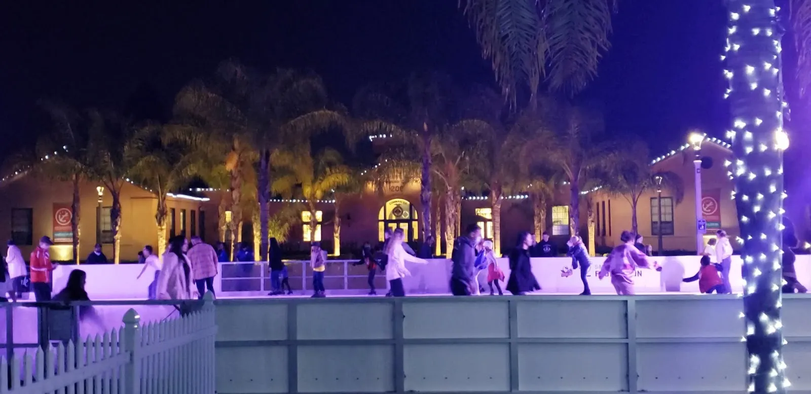 La pista de noche en la plaza, con una cerca blanca de madera al frente y las arcadas iluminadas y las palmeras de Liberty Station detrás. — Rady Children's Ice Rink