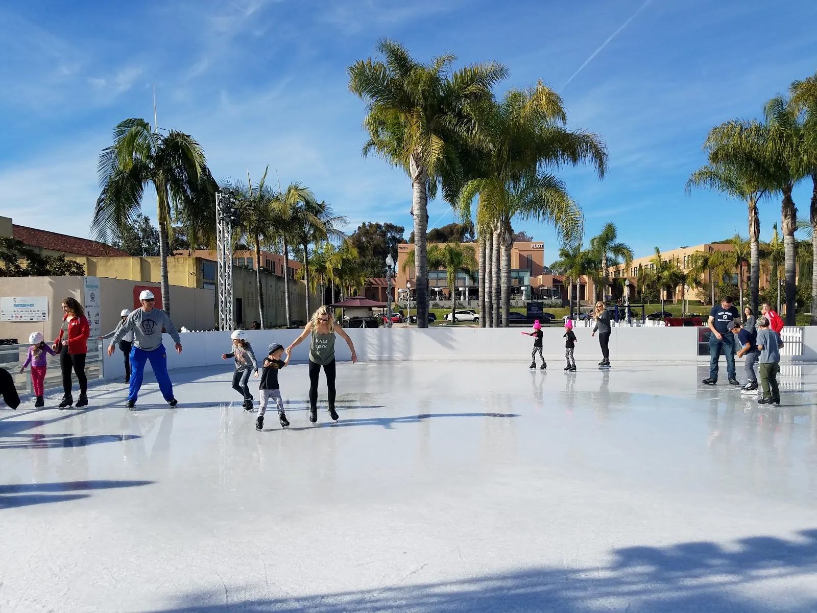La pista exterior de día bajo altas palmeras, con algunos patinadores repartidos y bandas blancas bajas alrededor del borde. — Rady Children's Ice Rink