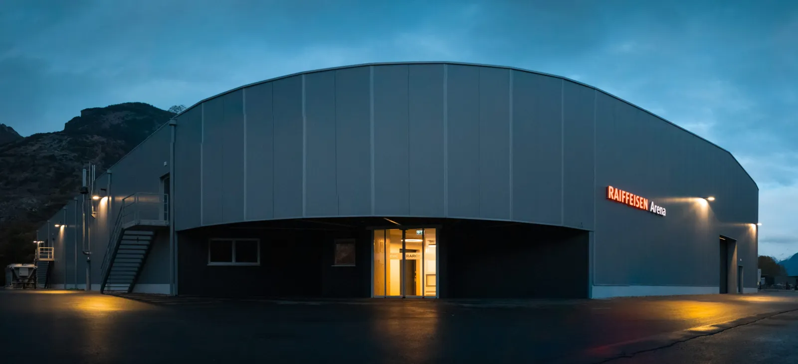 Die graue Raiffeisen Arena in Raron in der Dämmerung, von vorne gesehen mit beleuchtetem Eingang – Raiffeisen Arena Raron