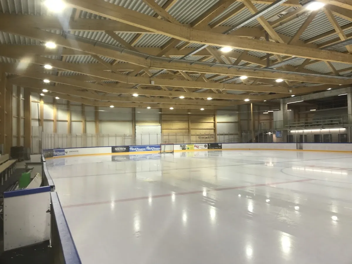 Die leere Eisfläche der Raiffeisen Arena unter ihren hölzernen Dachbindern, von den Banden aus gesehen – Raiffeisen Arena Raron