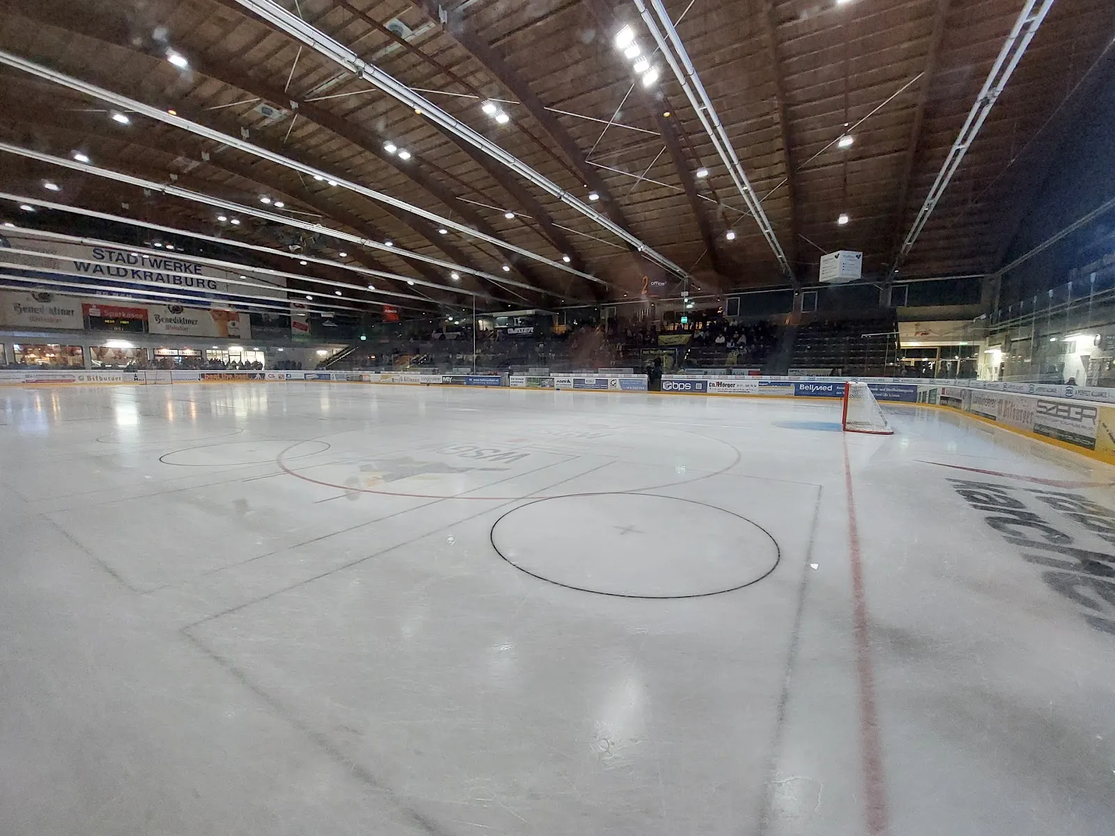 The empty ice sheet seen at rink level with faded face-off circles, stand boards along the far side and a timber roof with long strip lights overhead. — Raiffeisen Arena Waldkraiburg
