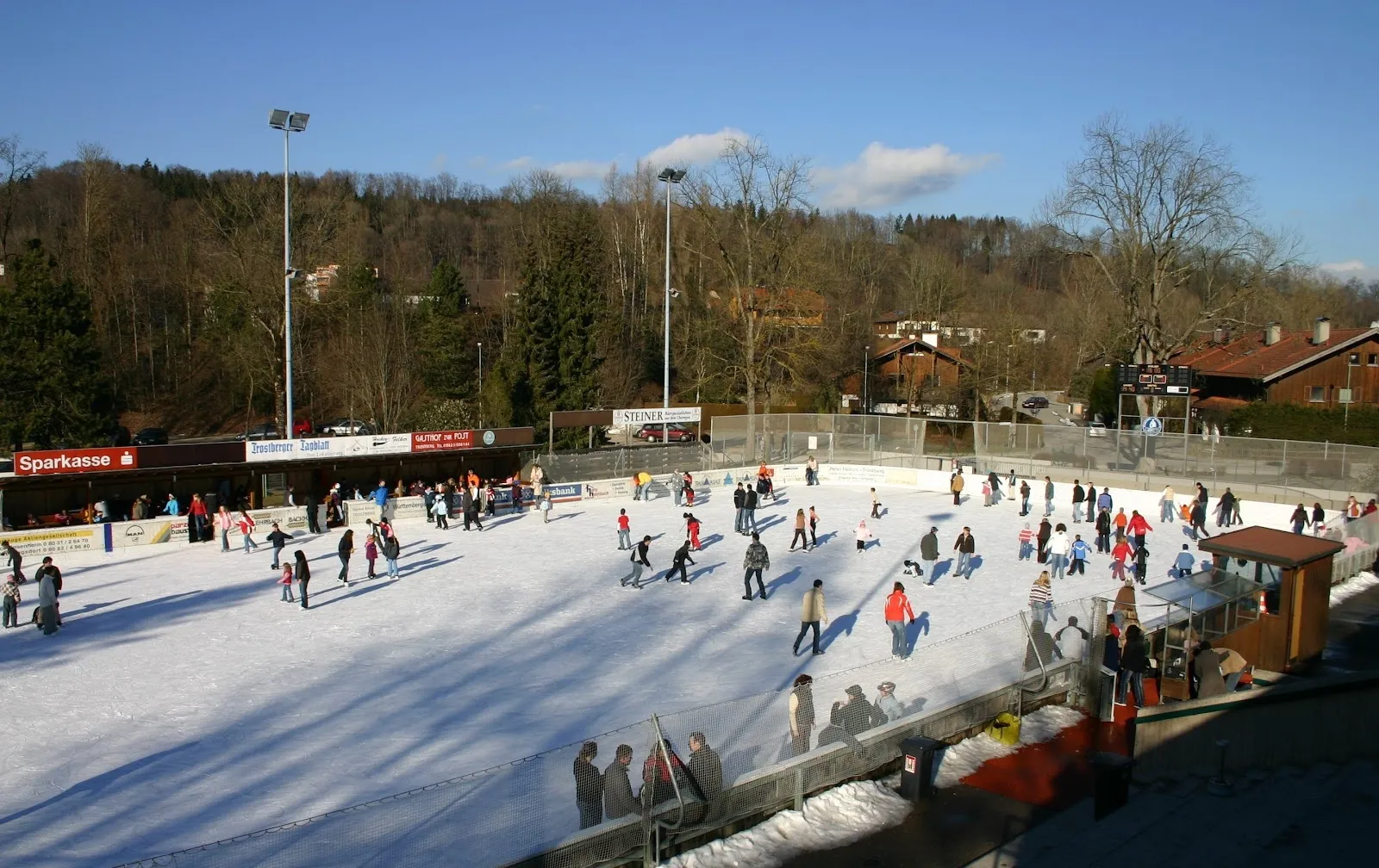 Das Freiluft-Rainer-Roßmanith-Eisstadion in Trostberg an einem Wintertag, mit Läufern auf der großen Fläche und dem bewaldeten Hang und dem Vereinsheim dahinter. – Rainer-Roßmanith-Eisstadion Trostberg