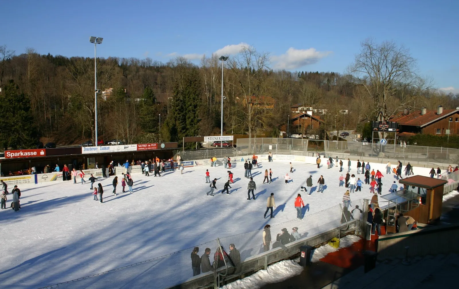 Die Freiluft-Eisbahn an einem sonnigen Wintertag, Läufer, die sich über das Eis verteilen, unter einem bewaldeten Hang, mit Flutlichtmasten und dem Vereinsheim an der gegenüberliegenden Seite. – Rainer-Roßmanith-Eisstadion Trostberg