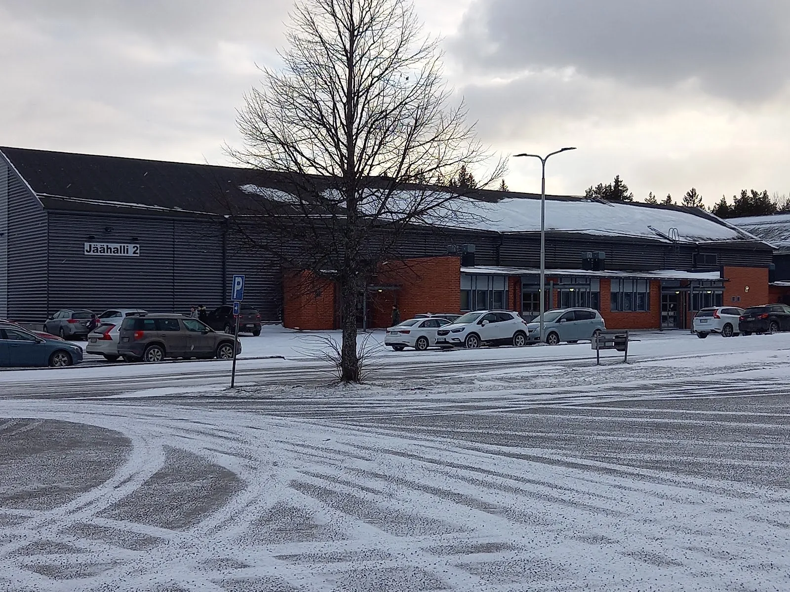 Raision jäähallien etuosa Kerttulassa talvella: tummanharmaa teräshalli tiilisine sisäänkäyntisiipineen ja Jäähalli 2 -kyltti, nähtynä lumisen parkkipaikan yli, edessä paljas puu – Raision jäähallit