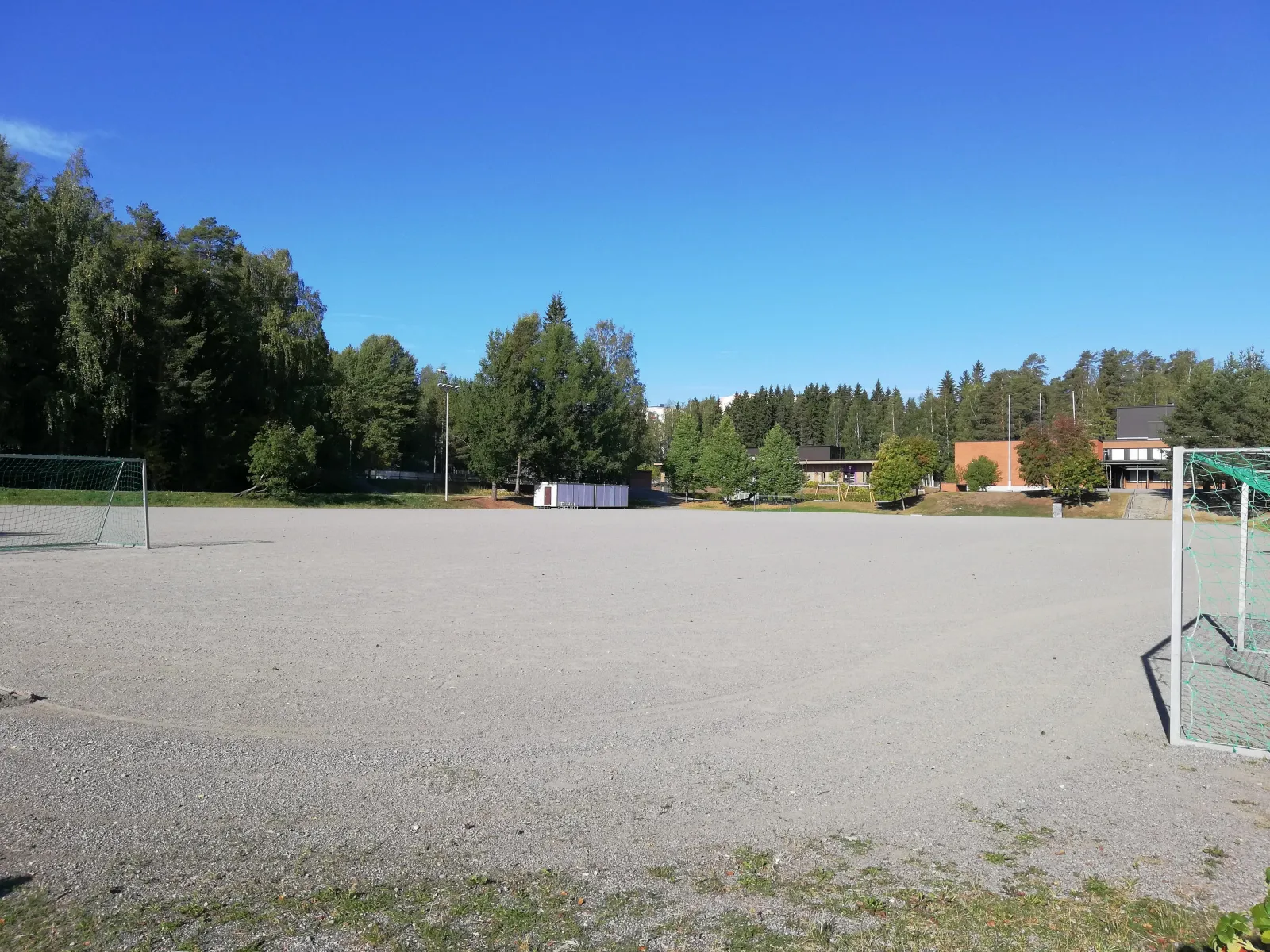 The wide gravel field at Rajala in Kuopio, which serves as a skating field in winter, seen in summer with a goal at the right edge and woods behind. — Rajalan luistelukenttä