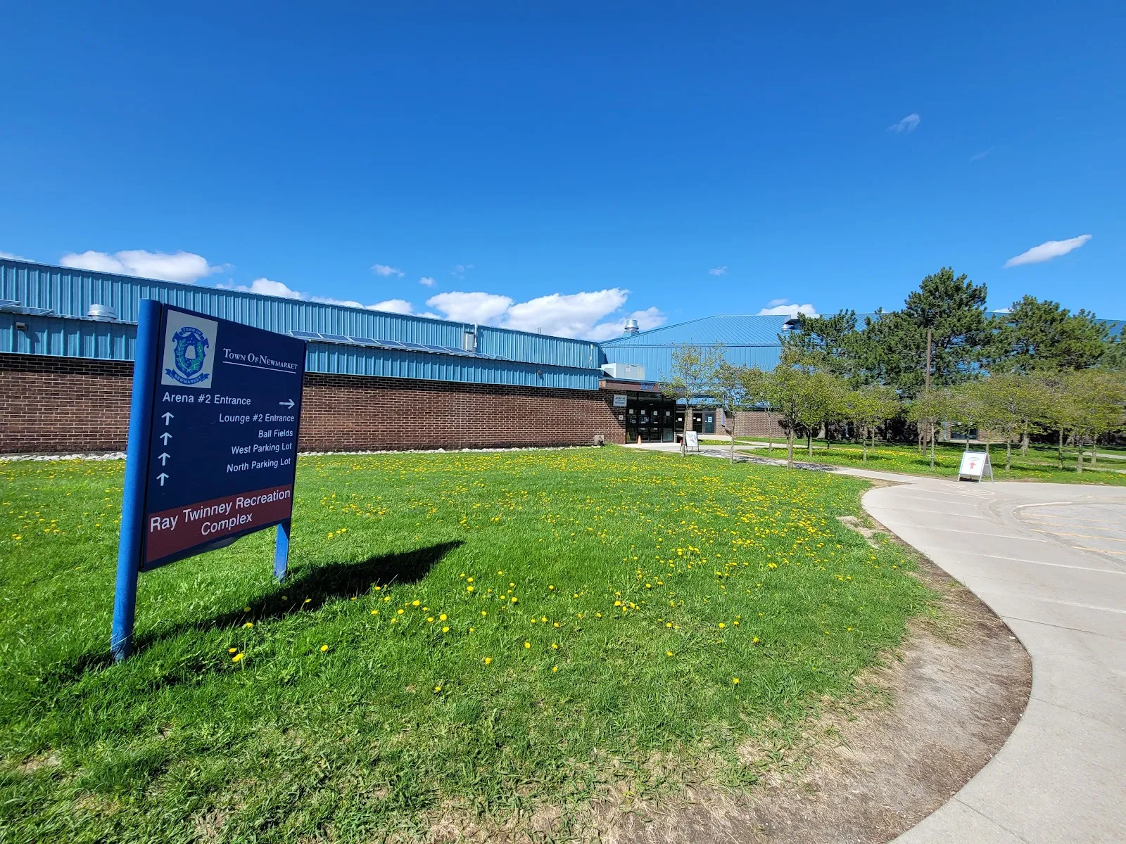 The Ray Twinney Recreation Complex sign on the lawn in front of the brick building with its blue metal roofline. — Ray Twinney Recreation Complex