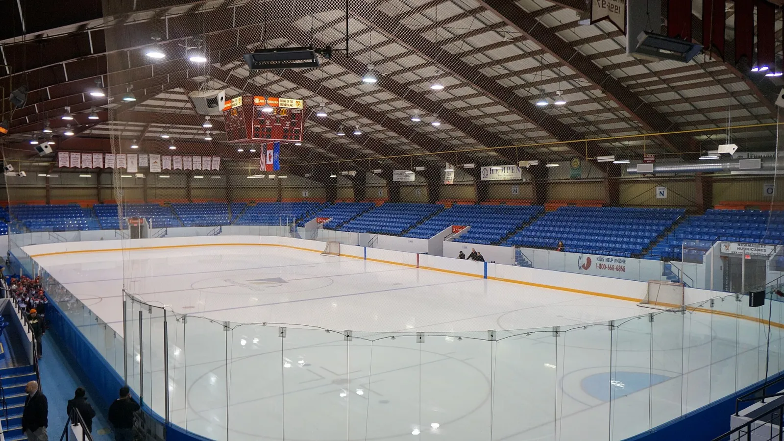 The main arena’s empty rink from the stands, with blue seats around the ice under a dark truss roof. — Ray Twinney Recreation Complex
