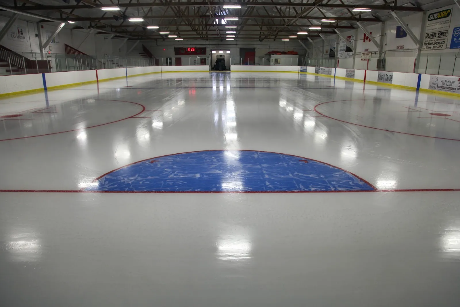 The empty ice of the Raymond Ice Arena with the blue crease, red lines and the boards down the length of the rink — Raymond Ice Arena