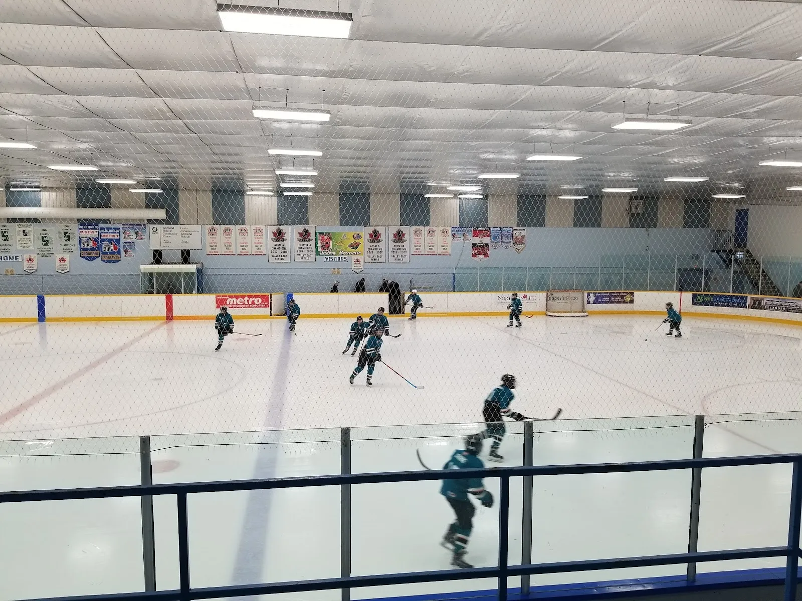 Un partido visto por encima del vidrio de una pista amplia, con jugadores de verde azulado repartidos por el hielo bajo un techo blanco acanalado del que cuelgan banderines de equipos. — Raymond Plourde Arena (Val Caron)