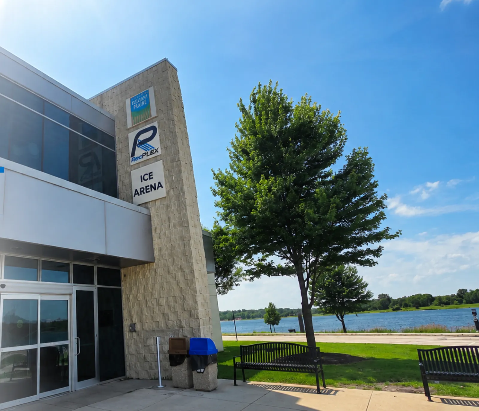 The building entrance, a tall stone-clad tower with stacked "Pleasant Prairie", "RecPlex" and "ICE ARENA" signs, glass doors below and a lake with a walking path across the parking lot. — RecPlex Ice Arena