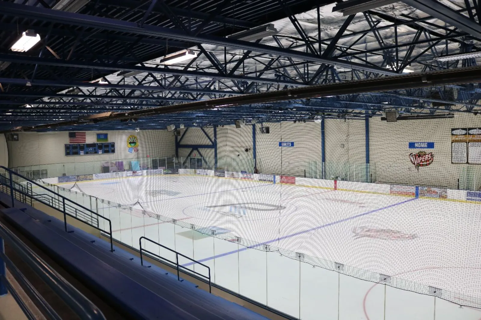 An empty NHL-sized ice sheet seen from the stands, with a blue steel-truss ceiling, a scoreboard and American flag on the end wall, and "HOME VIPERS" and "GUESTS" team-bench signage on the side boards. — RecPlex Ice Arena