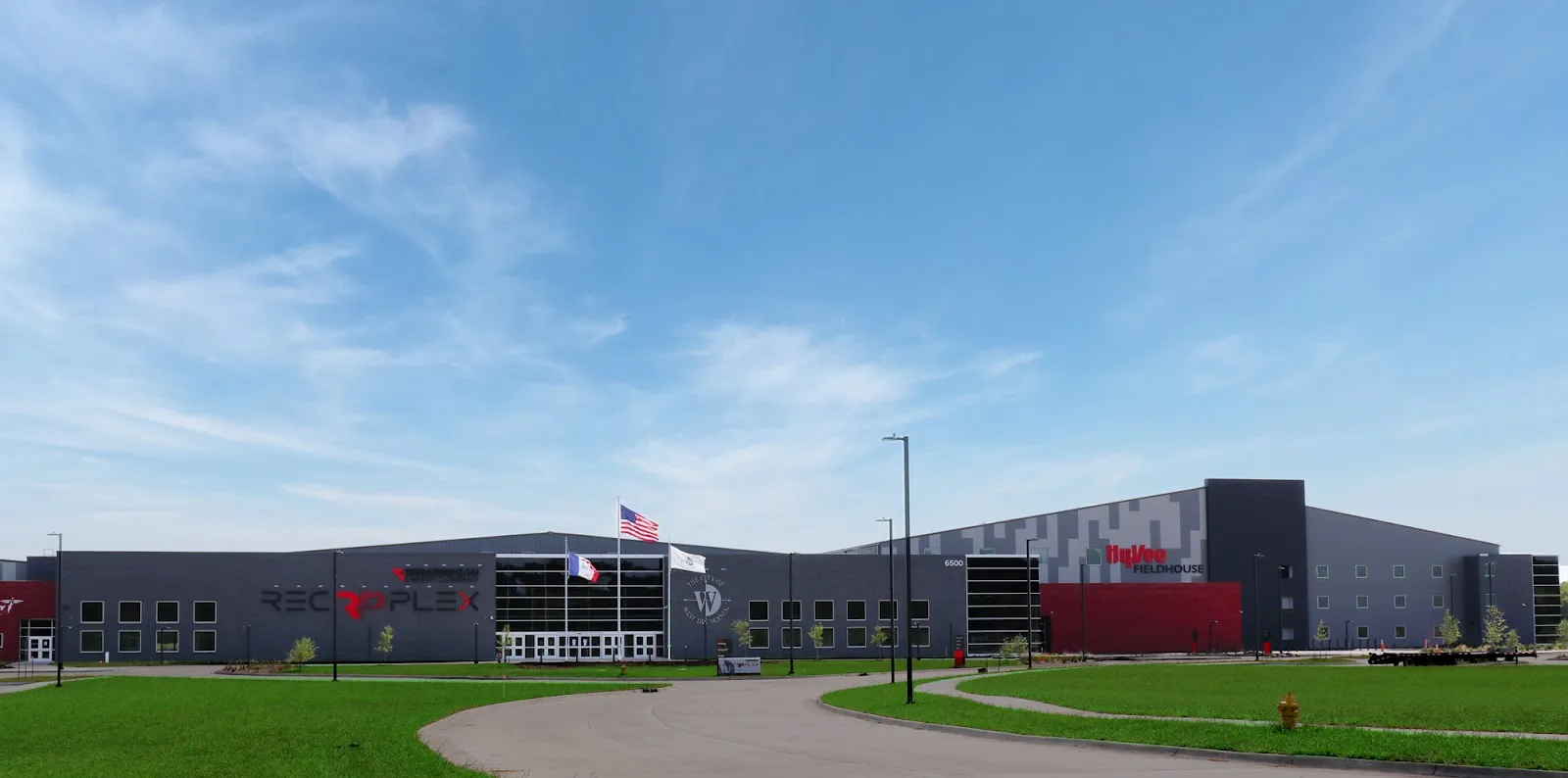 Front of The MidAmerican Energy Company RecPlex on a clear day, gray metal-panel building with a large red "RECPLEX" wordmark, American, Iowa and City of West Des Moines flags on poles, the connected HyVee Fieldhouse visible to the right, curved entry walkway and fresh landscaping. — MidAmerican Energy Company RecPlex