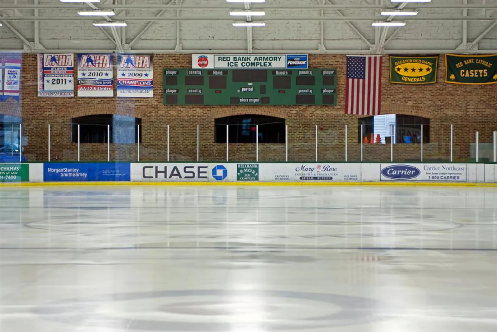 Toda la pista vacía bajo el techo en arco de ladrillo y acero, con un marcador verde que dice "RED BANK ARMORY ICE COMPLEX", una bandera de Estados Unidos, mantas de campeonato y anuncios de patrocinadores en las vallas; no hay personas en el hielo. — Red Bank Armory Ice Complex