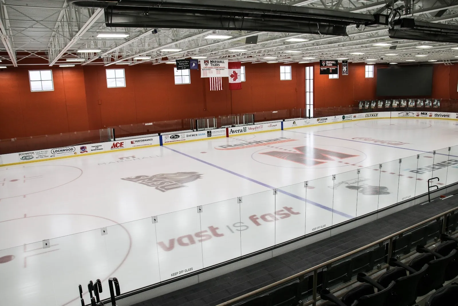 El hielo vacío bajo un alto techo con una pared de ladrillo rojo detrás, con el logotipo central de la arena y banderas de Canadá y Estados Unidos sobre las vallas del extremo. — Red Baron Arena & Expo