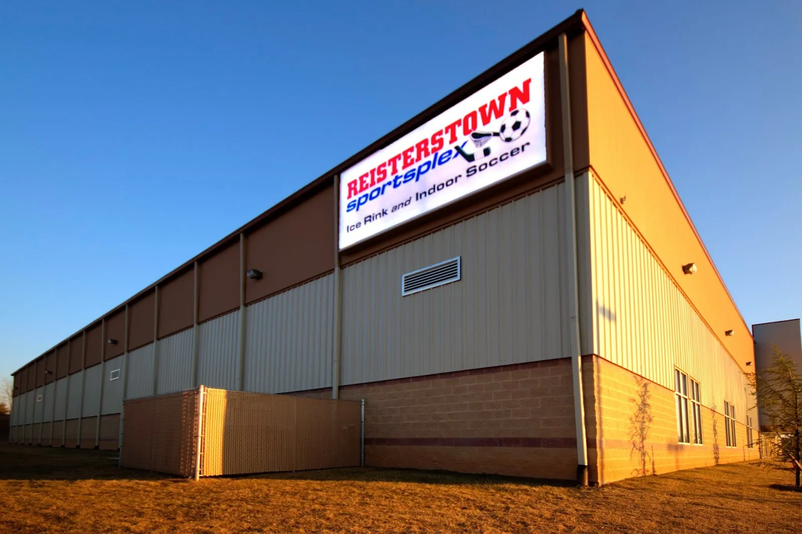 Corner of the tan metal-sided Reisterstown Sportsplex building at dusk, lit sign reading REISTERSTOWN sportsplex / Ice Rink and Indoor Soccer mounted high on the wall, chain-link enclosure and young trees in front. — Reisterstown Sportsplex