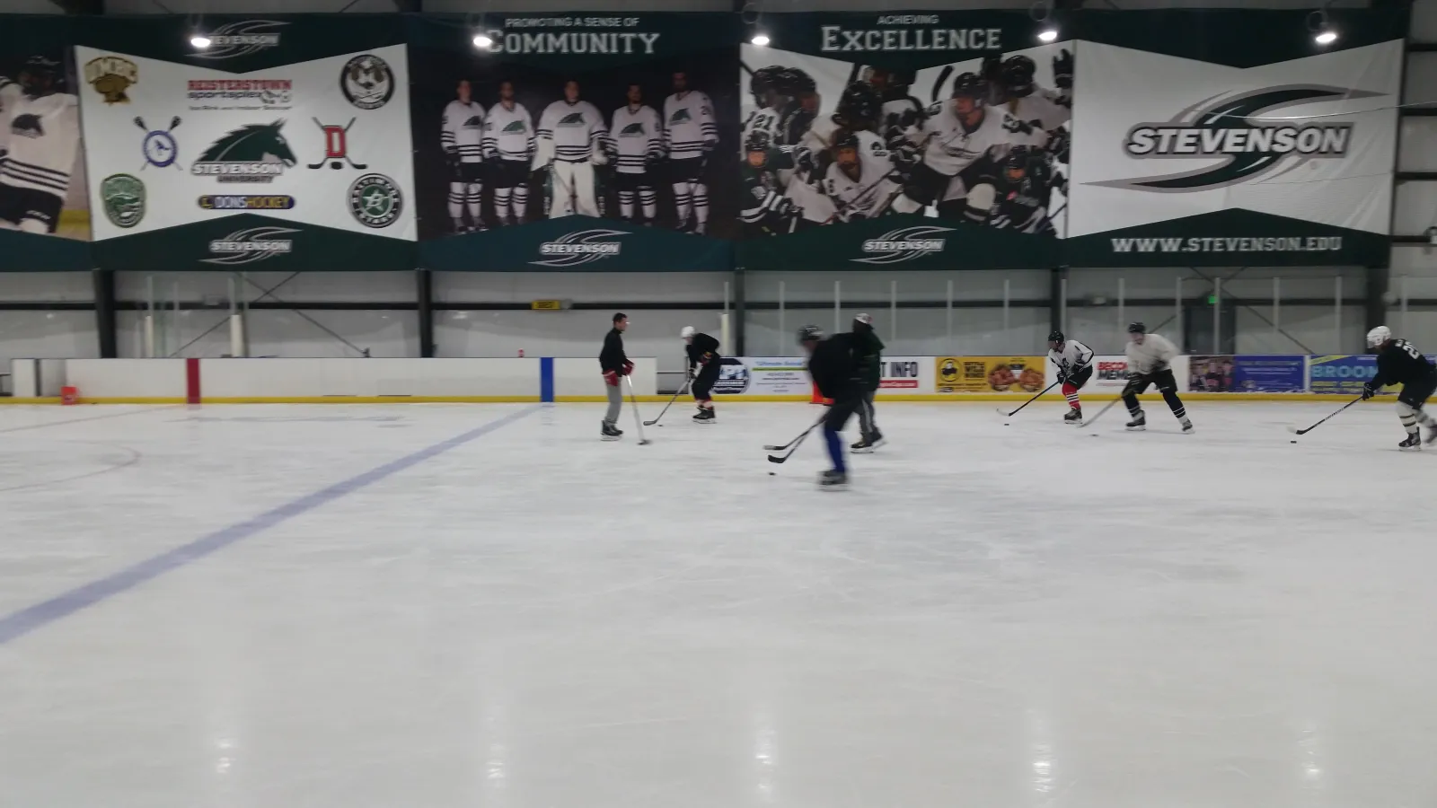 Wide view of the ice sheet during a stick-and-puck session, several skaters with hockey sticks spread across the rink, a small 'REISTERSTOWN sportsplex Ice Rink and Indoor Soccer' banner and Stevenson University hockey banners mounted on the boards behind. — Reisterstown Sportsplex
