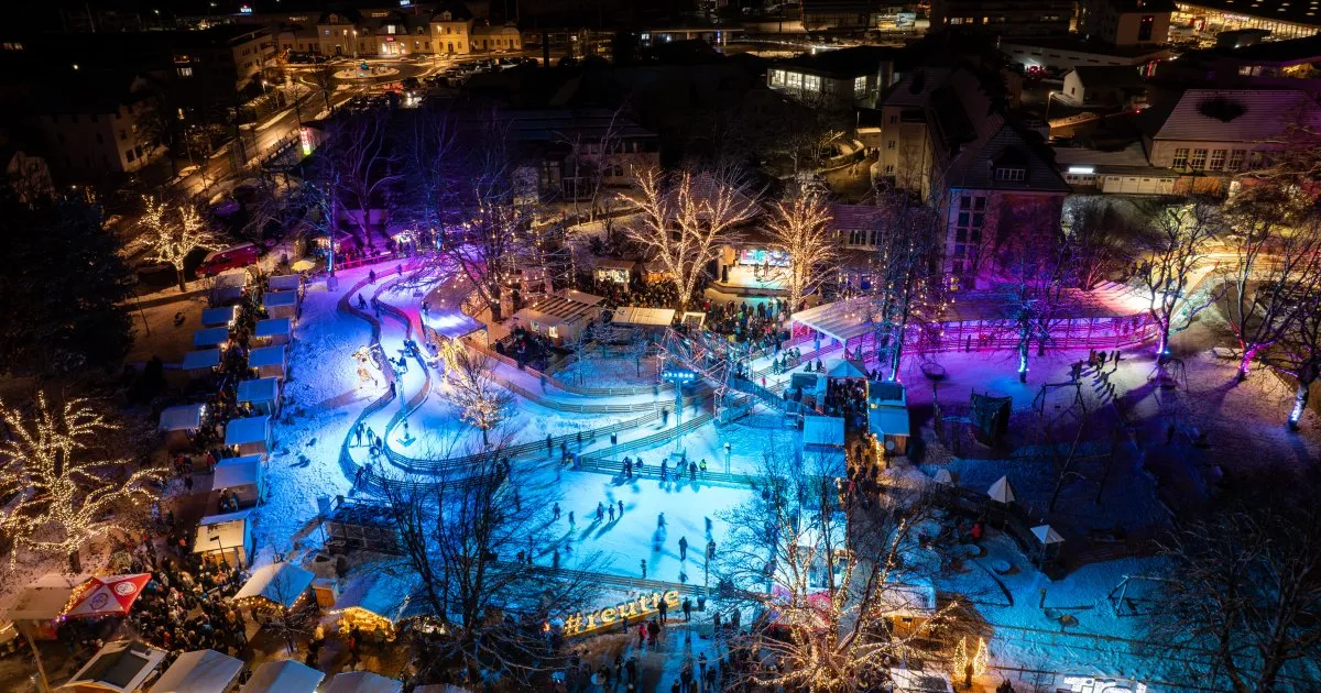 Nächtliche Drohnenaufnahme der Eisbahn Reutte on Ice im Stadtzentrum: die beleuchtete Eisfläche zwischen den Marktständen und den umliegenden Gebäuden; Eisläufer sind klein in der Ferne zu sehen. – Reutte on Ice
