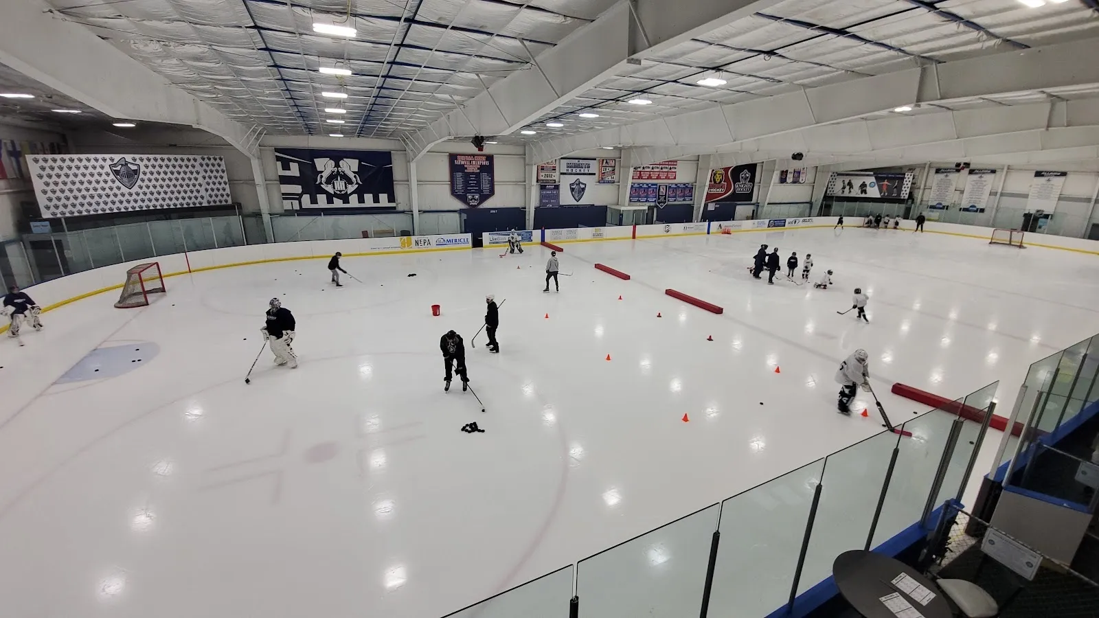 Una vista amplia desde el extremo de la pista, con unos cuantos jugadores repartidos por el hielo bajo un techo claro con banderines colgados a lo largo de la pared del fondo. — Revolution Ice Centre