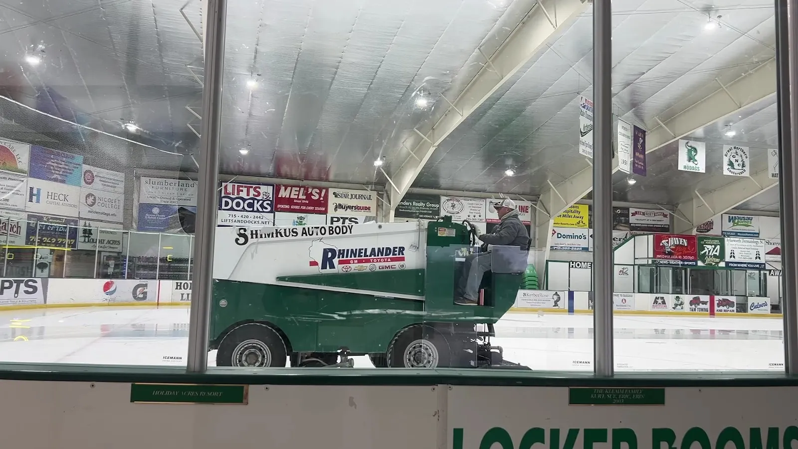 A green Zamboni marked Rhinelander resurfacing the ice, seen through the glass beneath rows of sponsor banners. — Rhinelander Ice Arena