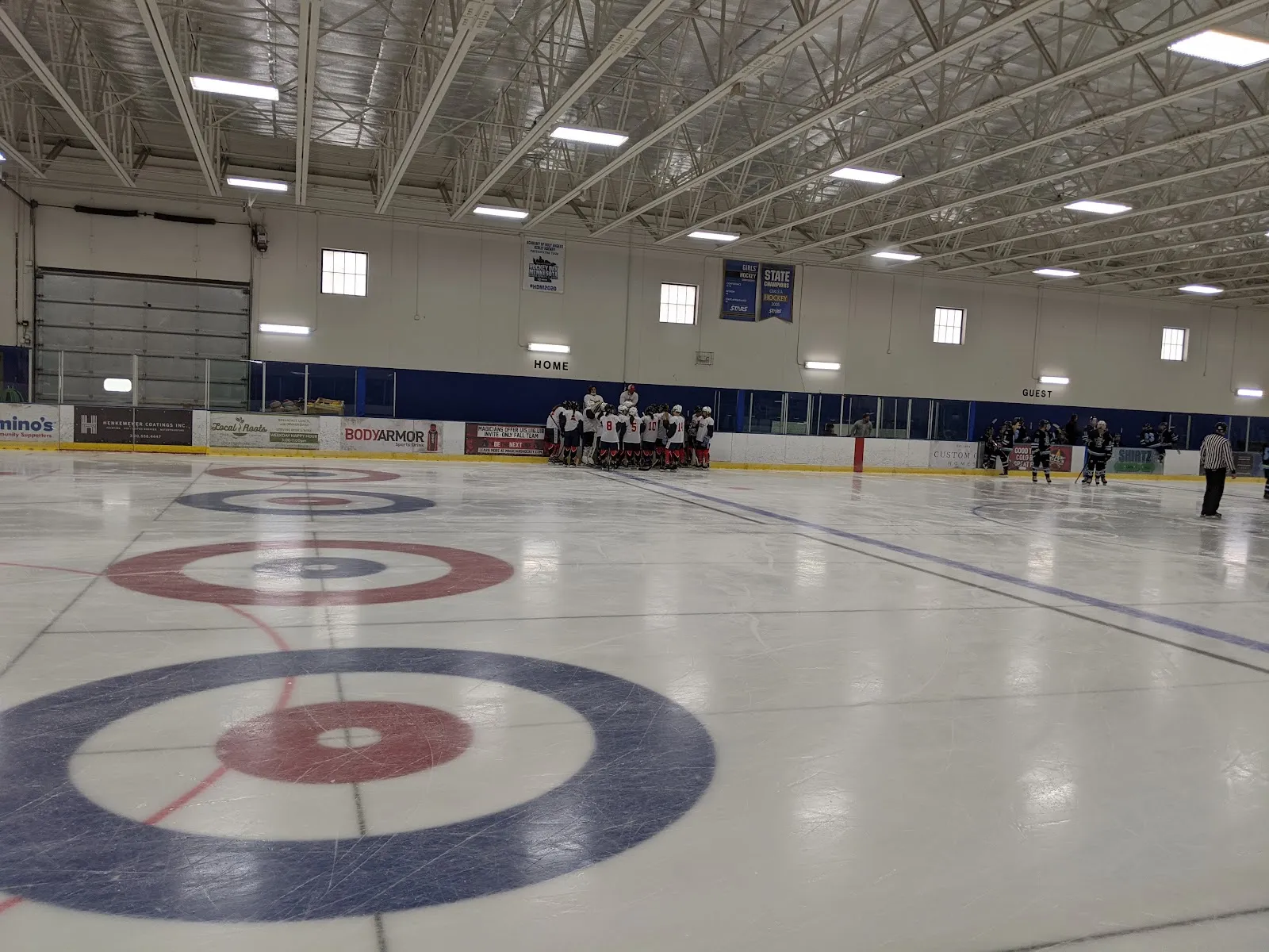 Interior of one ice sheet with "HOME"/"GUEST" wall lettering, retired-number and state-tournament banners, and an Academy of Holy Angels hockey banner on the far wall; a curling target is painted on the near ice and a youth team huddles at the boards. — Richfield Ice Arena
