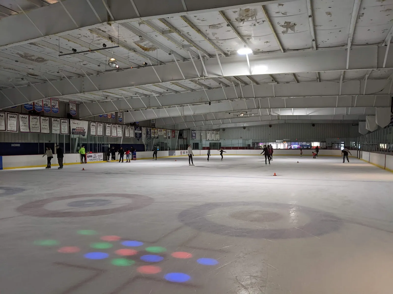 Interior of the rink from ice level: full sheet visible with curling-style target markings on the ice, a handful of public skaters spread across the surface, team championship banners along the boards, and the arched white steel roof overhead. — Richmond Ice Zone (North Chesterfield)